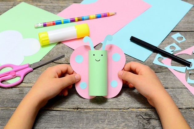 A child’s hands holding a paper butterfly over a table, with several sheets of paper, a pair of scissors, a glue stick and pencil on the table